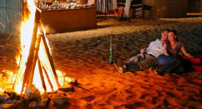 A couple sits on the sand in Qatar, leaning against each other and smiling by a campfire at night—one of the many romantic outdoor experiences. A hookah rests beside them, with colorful market stalls glowing in the background.