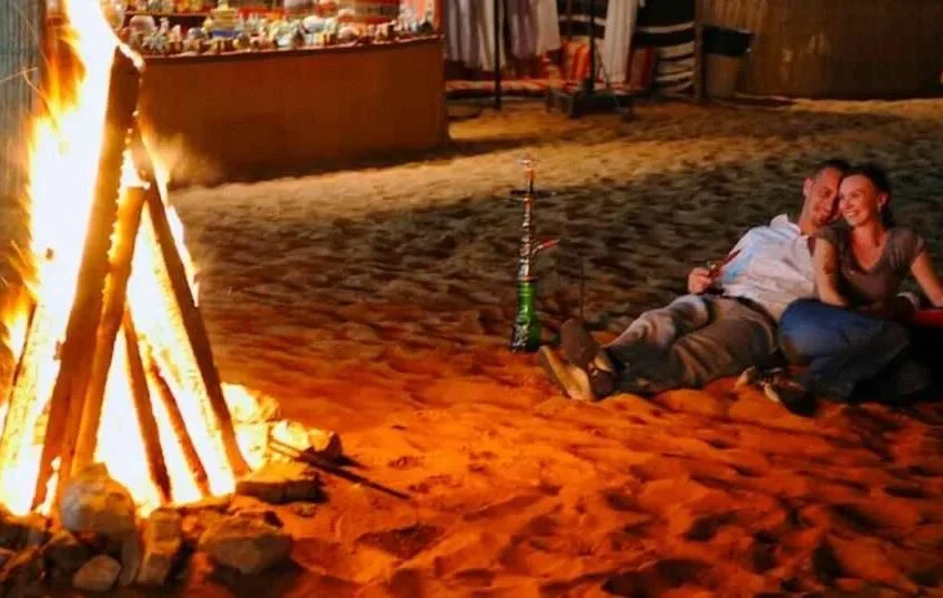 A couple sits on the sand in Qatar, leaning against each other and smiling by a campfire at night—one of the many romantic outdoor experiences. A hookah rests beside them, with colorful market stalls glowing in the background.