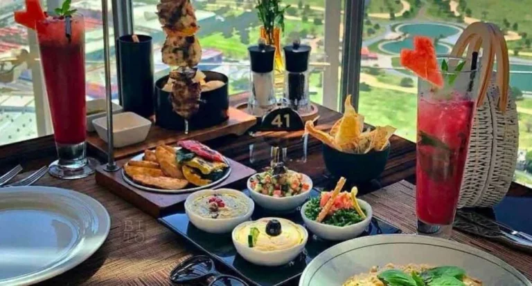 A table set with assorted dishes, dips, bread, grilled skewers, fries, and two tall red drinks garnished with watermelon slices, overlooking a scenic view through a large window at one of the best dining spots in Doha.