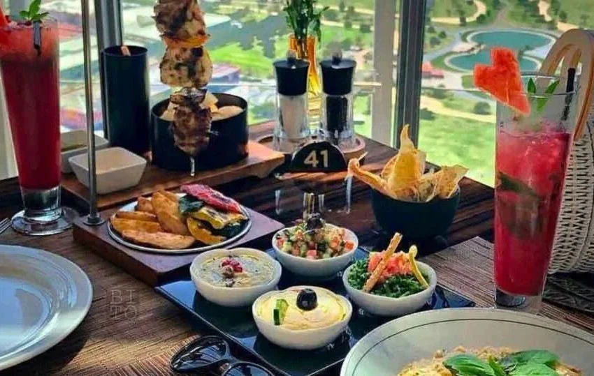A table set with assorted dishes, dips, bread, grilled skewers, fries, and two tall red drinks garnished with watermelon slices, overlooking a scenic view through a large window at one of the best dining spots in Doha.