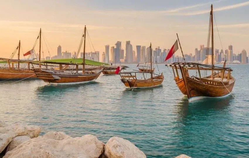 Traditional wooden boats float on calm blue water near a rocky shore, with a modern city skyline and tall skyscrapers in the background under a golden sky—capturing one of the best things to do in Doha.
