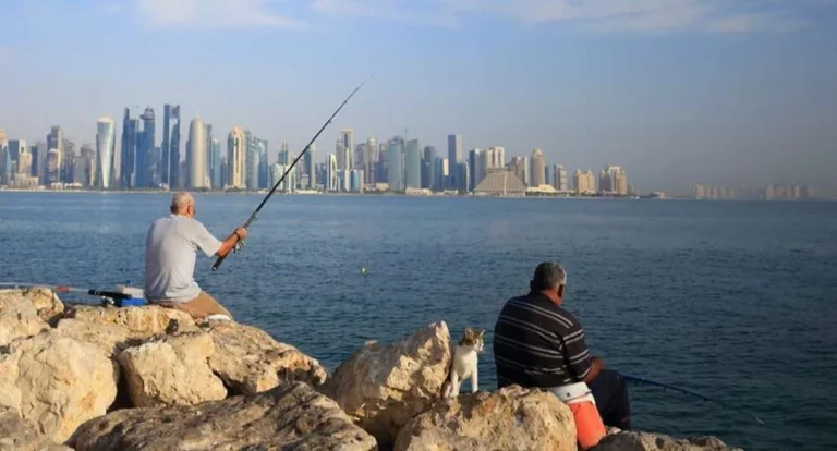 Two men sit on rocks fishing by the waterfront, enjoying one of the popular fishing spots in Qatar, with a city skyline visible across the water under a blue sky.