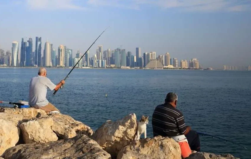 Two men sit on rocks fishing by the waterfront, enjoying one of the popular fishing spots in Qatar, with a city skyline visible across the water under a blue sky.
