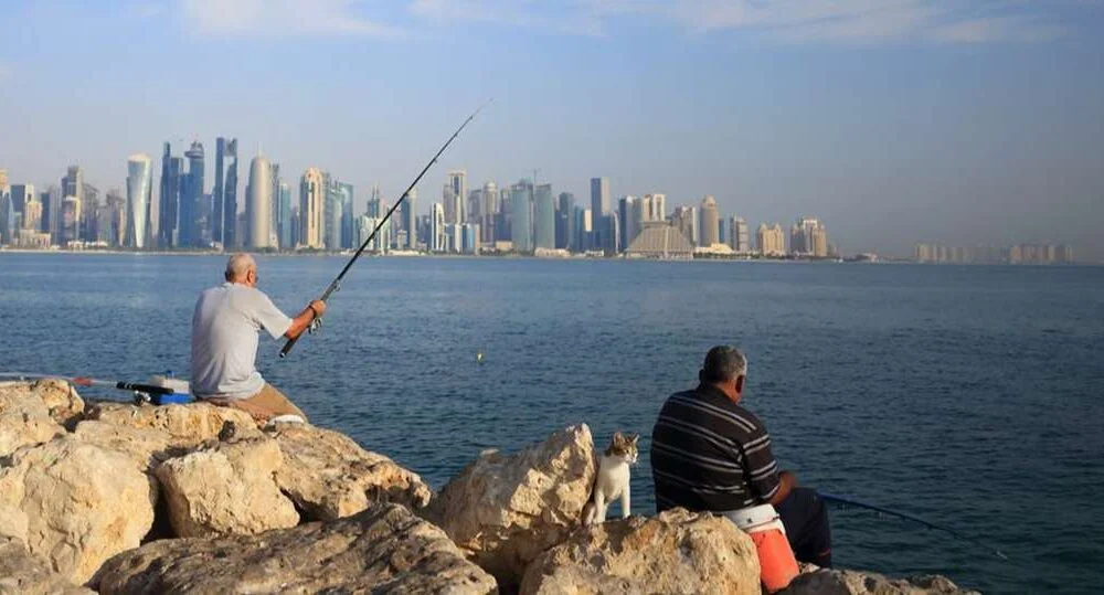 Two men sit on rocks fishing by the waterfront, enjoying one of the popular fishing spots in Qatar, with a city skyline visible across the water under a blue sky.