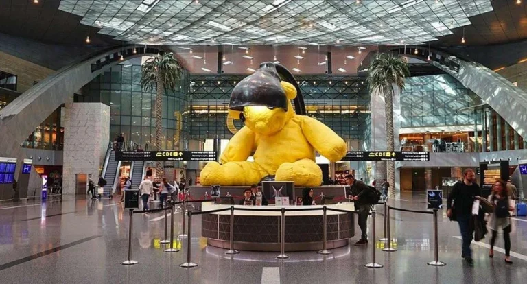 A large yellow teddy bear sculpture with a black lamp on its head greets travelers in the center of a modern airport terminal—remembering key airport tips can make your journey through such bustling spaces smoother.