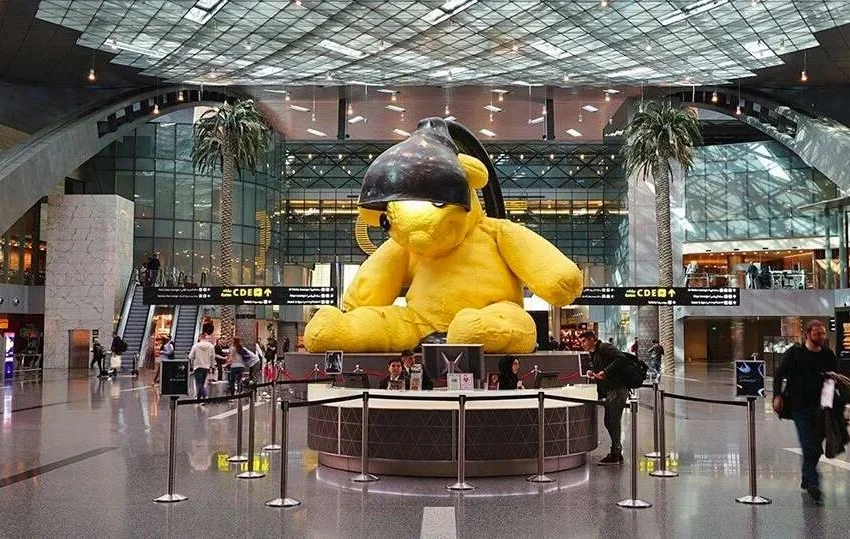A large yellow teddy bear sculpture with a black lamp on its head greets travelers in the center of a modern airport terminal—remembering key airport tips can make your journey through such bustling spaces smoother.