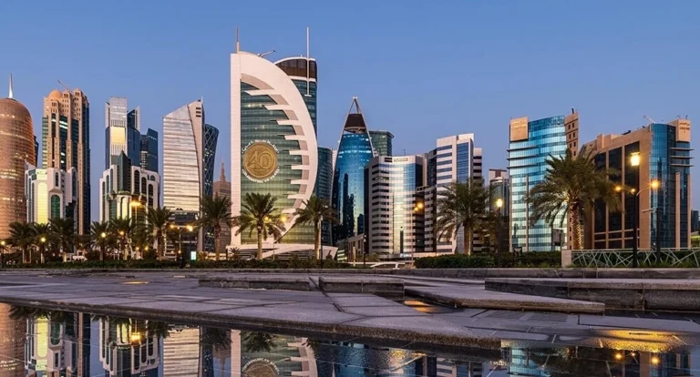 Modern city skyline with distinctive high-rise buildings and palm trees reflected in a body of water at dusk, against a clear blue sky—capturing the vibrant essence of one day in Doha.