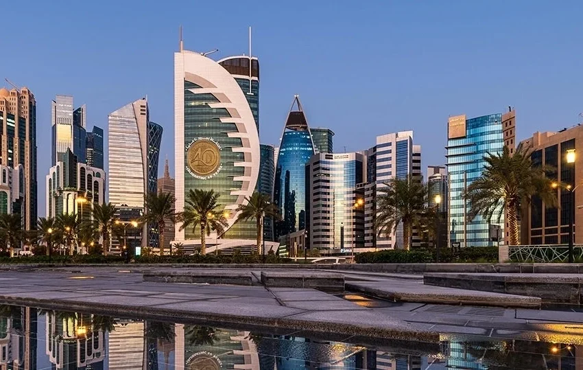 Modern city skyline with distinctive high-rise buildings and palm trees reflected in a body of water at dusk, against a clear blue sky—capturing the vibrant essence of one day in Doha.