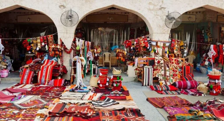 A vibrant market stall displays colorful woven rugs, blankets, and pillows—some of the best gifts to buy in Doha—with intricate patterns, set before arched stone doorways. Fans are mounted on the walls above the stalls.