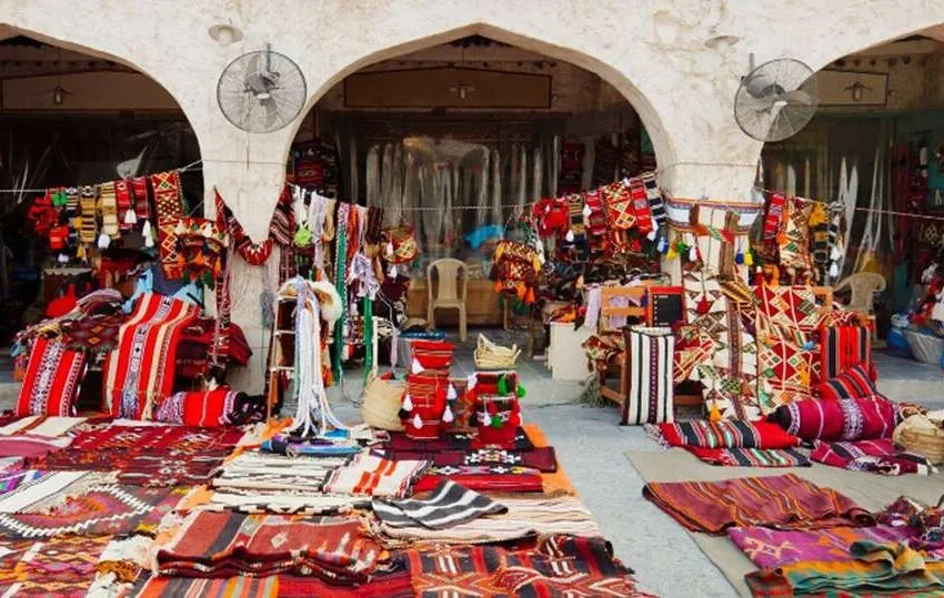 A vibrant market stall displays colorful woven rugs, blankets, and pillows—some of the best gifts to buy in Doha—with intricate patterns, set before arched stone doorways. Fans are mounted on the walls above the stalls.