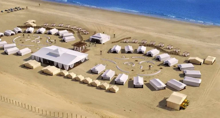 Aerial view of a luxury desert campsite on a sandy beach, featuring large white tents arranged in curved patterns with desert camping essentials, cabanas near the water, and pathways with small groups of people exploring. Blue ocean in the background.
