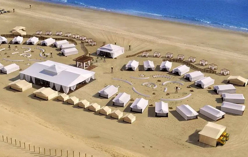 Aerial view of a luxury desert campsite on a sandy beach, featuring large white tents arranged in curved patterns with desert camping essentials, cabanas near the water, and pathways with small groups of people exploring. Blue ocean in the background.