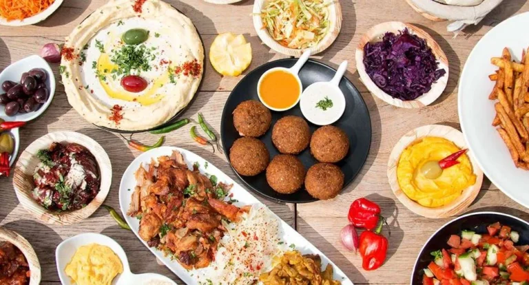 A variety of Middle Eastern dishes and traditional Qatari food on a wooden table, including hummus, falafel, rice with grilled meat, colorful salads, french fries, olives, dips, and fresh vegetables.