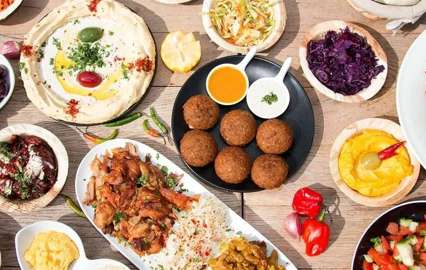 A variety of Middle Eastern dishes and traditional Qatari food on a wooden table, including hummus, falafel, rice with grilled meat, colorful salads, french fries, olives, dips, and fresh vegetables.