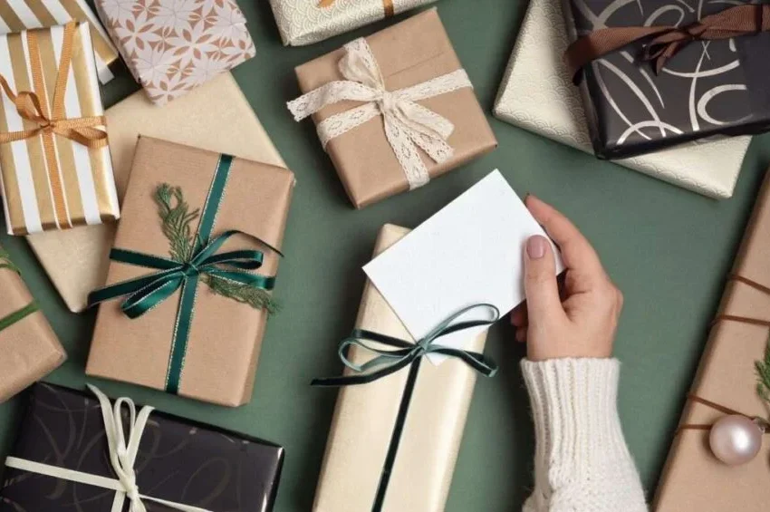 A hand in a cream sweater holds a blank card among neatly wrapped Ramadan Gifts with ribbons and holiday decorations, all arranged on a green surface.