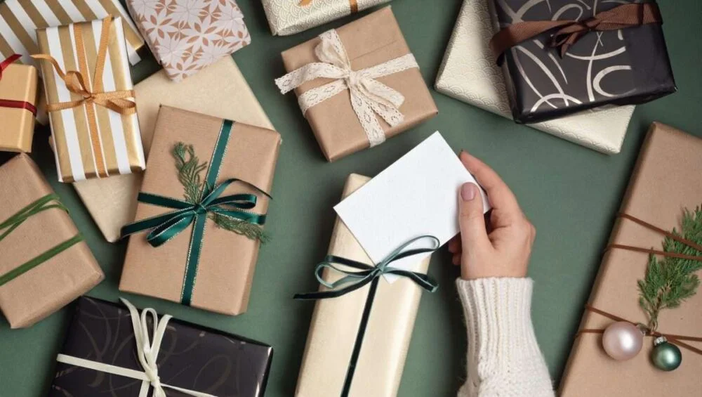 A hand in a cream sweater holds a blank card among neatly wrapped Ramadan Gifts with ribbons and holiday decorations, all arranged on a green surface.