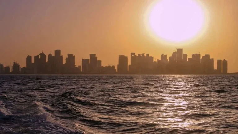 A city skyline silhouetted against a golden sunset, with the sun low in the sky and its light reflecting on rippling water—much like the breathtaking views enjoyed from some of the Best Restaurants in Doha.