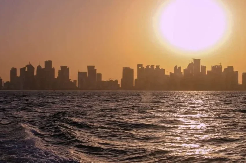 A city skyline silhouetted against a golden sunset, with the sun low in the sky and its light reflecting on rippling water—much like the breathtaking views enjoyed from some of the Best Restaurants in Doha.