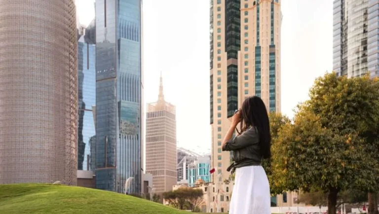 A woman in a white dress stands on green grass, looking up at tall modern skyscrapers—an inspiring moment for any Qatar itinerary, with trees nearby and sunlight brightening the cityscape.