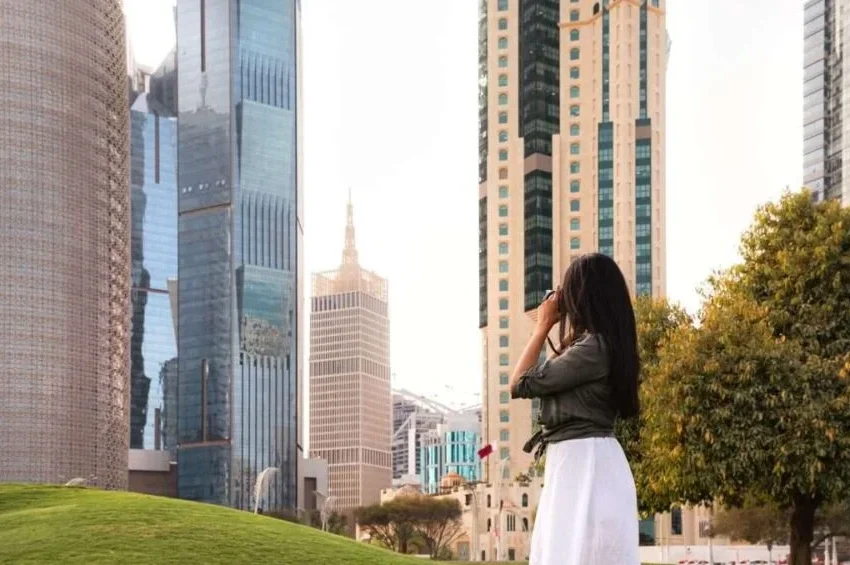 A woman in a white dress stands on green grass, looking up at tall modern skyscrapers—an inspiring moment for any Qatar itinerary, with trees nearby and sunlight brightening the cityscape.