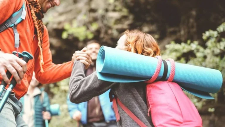 Two hikers greet each other with a handshake on a trail; one carries a blue rolled-up sleeping mat and a pink backpack. Reminiscent of outdoor adventures from the list of 10 Activities in Qatar, green foliage and rocks blur in the background.