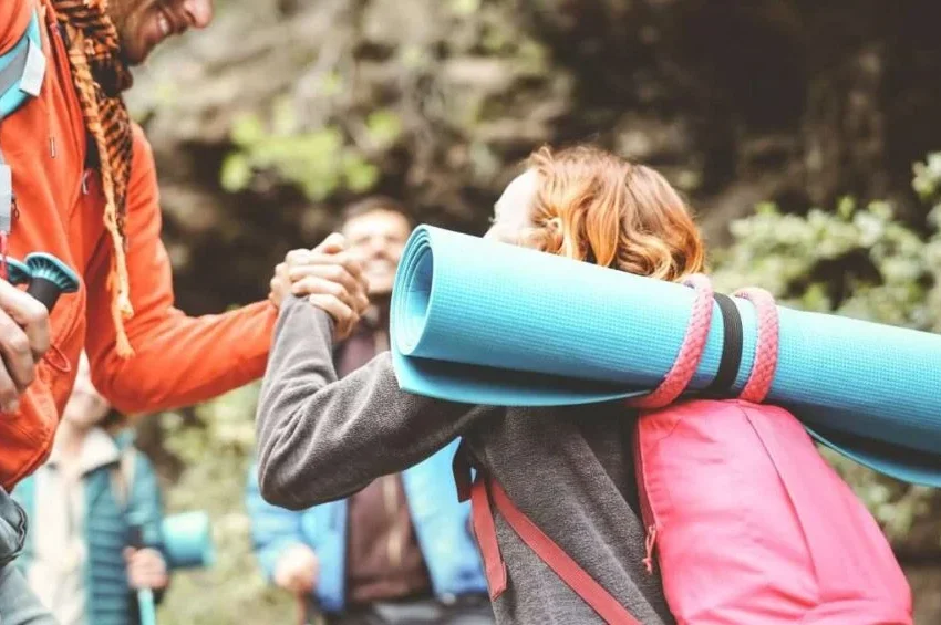 Two hikers greet each other with a handshake on a trail; one carries a blue rolled-up sleeping mat and a pink backpack. Reminiscent of outdoor adventures from the list of 10 Activities in Qatar, green foliage and rocks blur in the background.