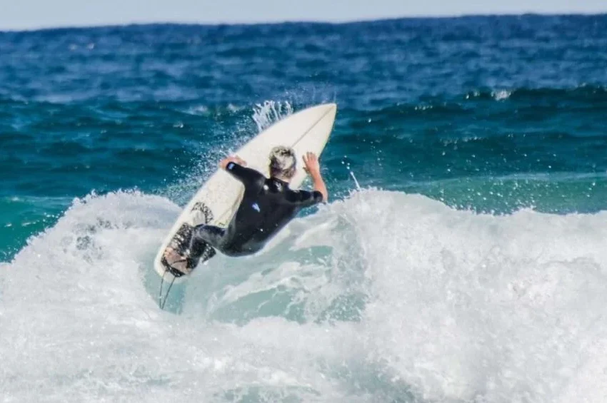 A surfer in a black wetsuit rides a wave, tilting their surfboard upward as white water splashes around them—capturing the excitement of water activities Qatar has to offer against the deep blue ocean and sky.