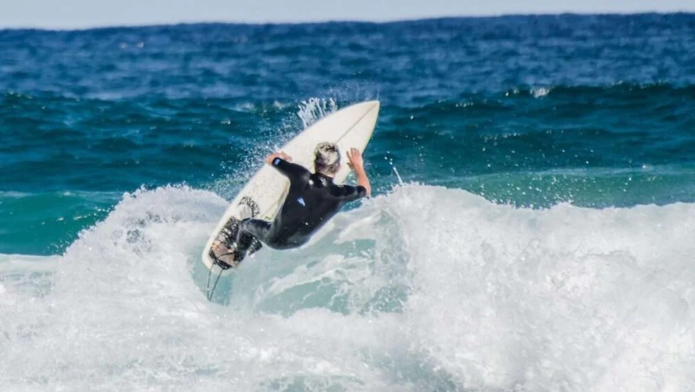 A surfer in a black wetsuit rides a wave, tilting their surfboard upward as white water splashes around them—capturing the excitement of water activities Qatar has to offer against the deep blue ocean and sky.