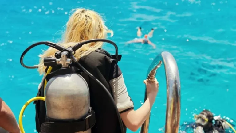 A scuba diver with blonde hair stands on a boat ladder, preparing to enter clear blue water for pearl diving in Qatar, while another person swims nearby and two divers are seen in the water below.