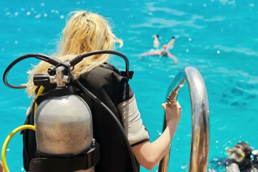 A scuba diver with blonde hair stands on a boat ladder, preparing to enter clear blue water for pearl diving in Qatar, while another person swims nearby and two divers are seen in the water below.