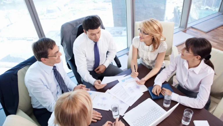 Five business professionals sit around a table in a modern office, discussing documents and charts related to the MICE Industry In Qatar. Laptops, papers, and glasses of water are on the table, with city buildings visible through large windows.