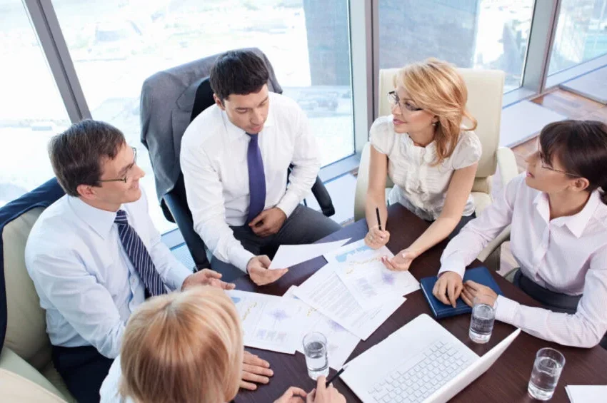 Five business professionals sit around a table in a modern office, discussing documents and charts related to the MICE Industry In Qatar. Laptops, papers, and glasses of water are on the table, with city buildings visible through large windows.