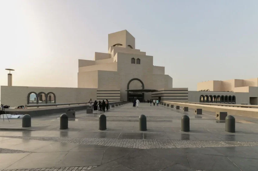 The image shows the exterior of Doha’s Museum of Islamic Art in Qatar, with its geometric, sand-colored stone architecture. A group of people walks toward the entrance on a wide pathway under a clear sky.