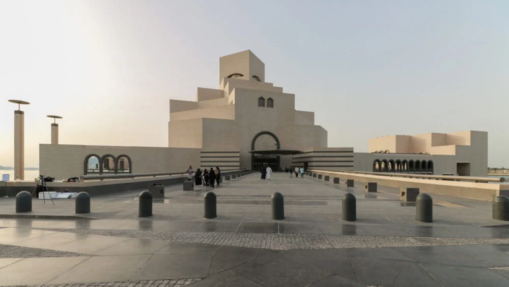 The image shows the exterior of Doha’s Museum of Islamic Art in Qatar, with its geometric, sand-colored stone architecture. A group of people walks toward the entrance on a wide pathway under a clear sky.