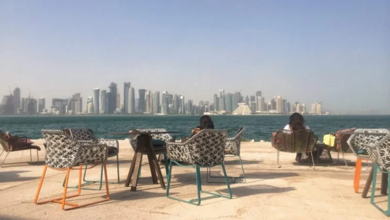 Outdoor seating area with patterned chairs and tables by the water, overlooking a modern city skyline under a clear sky. Two people are seated, enjoying one of the top tourist attractions in Qatar with stunning waterfront views.