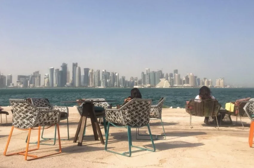 Outdoor seating area with patterned chairs and tables by the water, overlooking a modern city skyline under a clear sky. Two people are seated, enjoying one of the top tourist attractions in Qatar with stunning waterfront views.