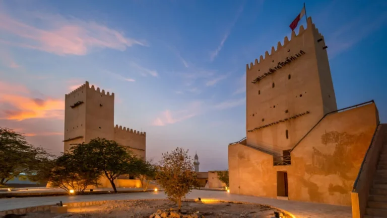 Two illuminated beige stone towers, known as Barzan Towers, stand against a colorful sunset sky in Qatar, their crenellated tops and fluttering flag marking them as notable historical landmarks amid trees and winding pathways.