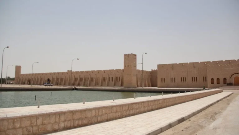 A large, beige stone fortress with tall walls and towers, reminiscent of the Sheikh Faisal Museum Qatar, stands next to a calm, man-made canal under a clear sky. Street lamps line the paved walkway beside the water.