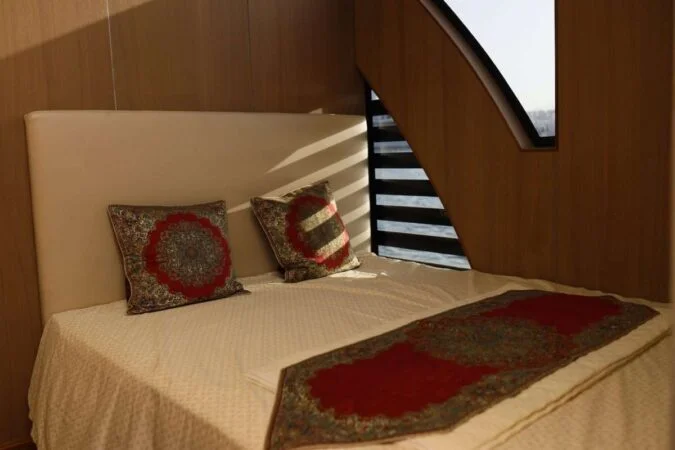 A neatly made bed with a cream-colored headboard, decorative pillows, and a patterned throw awaits you on your House Boat Tour Qatar. Sunlight pours in through windows with geometric black slats, casting serene shadows on the bed.