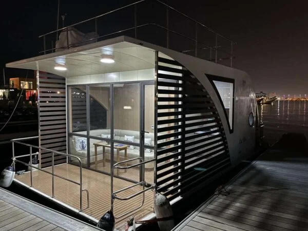Modern houseboat docked at night, illuminated from within. Large windows and horizontal slats adorn the sides, with a small deck and railing—perfect for a House Boat Tour Qatar—offering views of city lights reflecting on the water.
