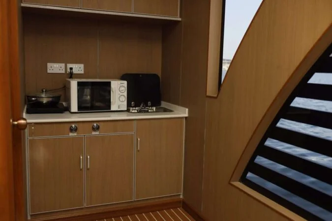 A compact kitchenette with brown cabinets, a white countertop, a microwave, and a gas stove sits next to a large window overlooking the water—perfect for enjoying views during your House Boat Tour Qatar adventure.
