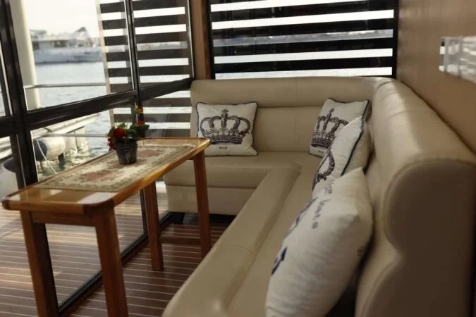 A cozy corner seating area with a cream-colored L-shaped sofa, crown-printed pillows, and a wooden table topped with a floral runner and small potted plant—perfect for relaxing during your House Boat Tour Qatar next to large windows overlooking the water.