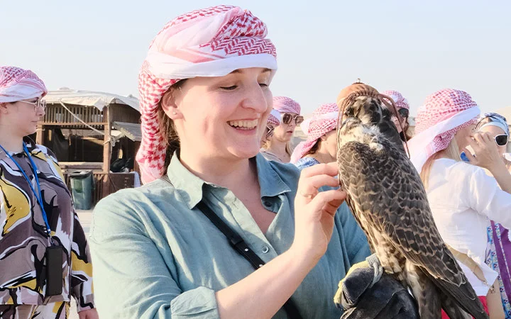 A smiling woman wearing a pink and white headscarf gently holds a falcon on her gloved hand during a Half Day Desert Safari Qatar. Several people in similar headscarves and sunglasses stand in the background at this outdoor event.