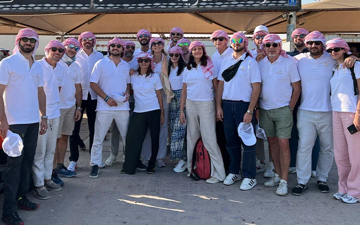 A group of people wearing white shirts, sunglasses, and red-and-white checkered headscarves pose together outdoors on a sunny day during a Half Day Desert Safari Qatar. Some hold hats, and a tent canopy is visible in the background.