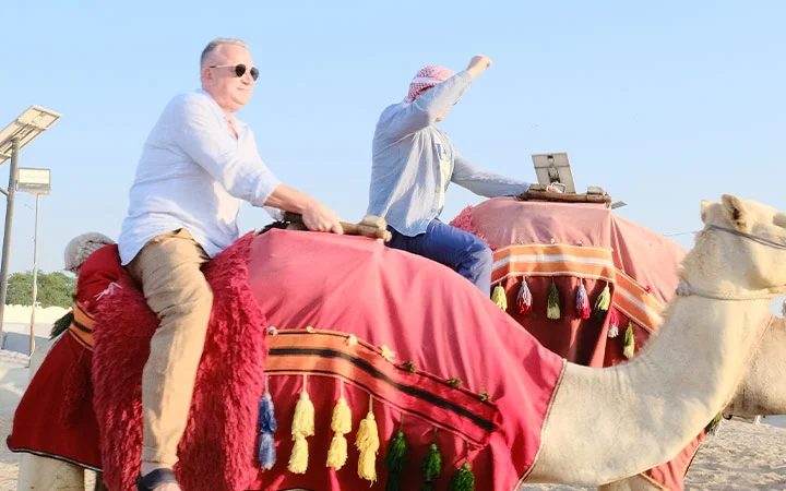 Two men ride camels draped in colorful, tasseled cloths beneath a clear blue sky. Perfect for a Half Day Desert Safari Qatar, the scene captures the excitement of exploring iconic desert landscapes with friends.