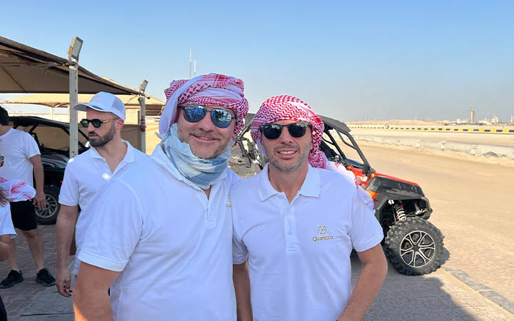 Two men in white polo shirts and sunglasses, wearing red-and-white keffiyehs, stand and smile outside near a dune buggy during a Half Day Desert Safari Qatar. Other men in white shirts and hats and a clear blue sky are in the background.