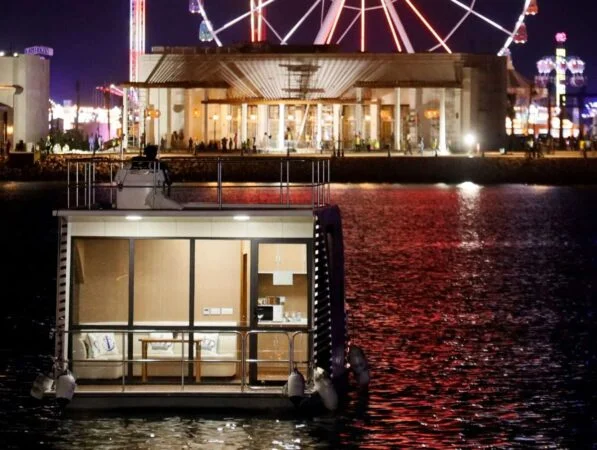 A small, illuminated houseboat floats on water at night during a House Boat Tour Qatar, with a large, brightly lit Ferris wheel and buildings reflecting in the background.