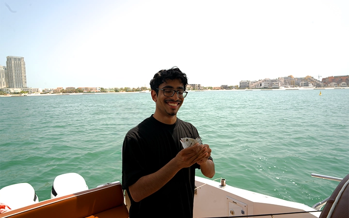 A smiling man wearing glasses holds a small fish while standing on a private speed boat fishing trip, with calm blue water and buildings visible in the background on a sunny day.