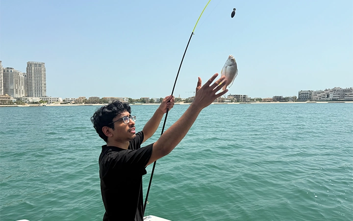 A young man on a Private Speed Boat Fishing Trip holds up a fishing rod with one hand while reaching for a fish he caught over the blue water, city buildings visible in the background under a clear sky.