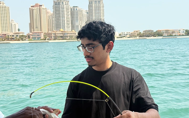 A person wearing glasses and a black shirt stands near the water, holding a yellow fishing rod during a Private Speed Boat Fishing Trip. Tall buildings and a scenic shoreline are visible in the background under a clear sky.